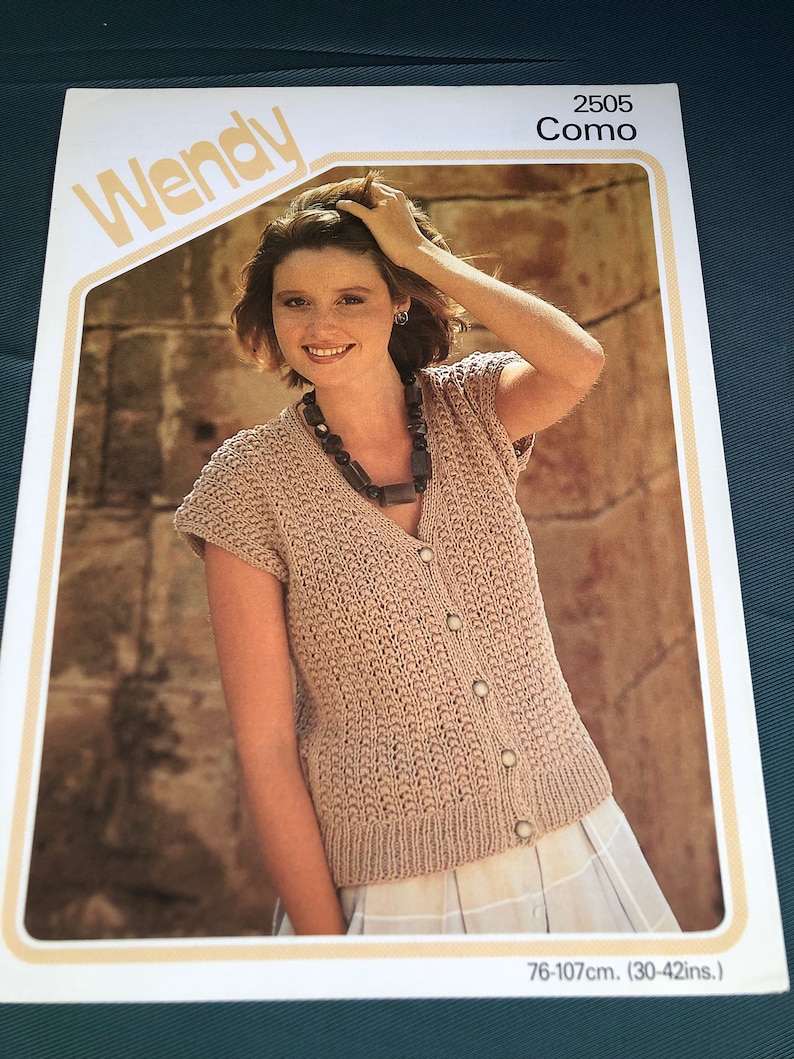 May include: A knitting pattern for a beige knitted cardigan with a V-neck and button closure. The pattern is called "Como" and is from Wendy, pattern number 2505. The pattern is for sizes 76-107 cm.