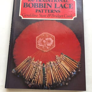 May include: A book titled "100 Traditional Bobbin Lace Patterns" by Geraldine Stott & Bridget Cook. The cover features a red background with a bobbin lace design and a collection of bobbins.
