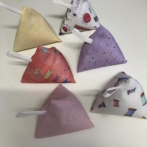 Könnte beinhalten: Sechs dreieckige Stoffsäckchen in verschiedenen Farben und Mustern, jedes mit einer weißen elastischen Schlaufe. Die Muster umfassen florale, nähbezogene und abstrakte Designs. Die Säckchen sind auf einer weißen Oberfläche angeordnet.