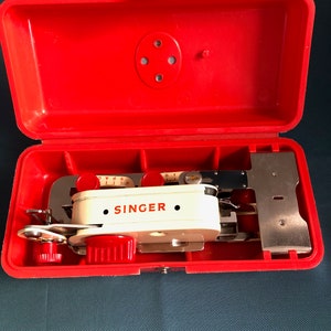 May include: A vintage Singer sewing machine attachment kit in a bright red plastic case. The kit includes a white and red sewing machine attachment with the Singer logo, along with various metal and plastic components. The case is open, revealing the contents.