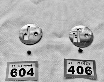 May include: Two circular metal parts with slots and holes, two small black pieces, and two stickers displaying the numbers 604 and 406. The items are on a white, textured surface.