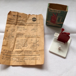 May include: A vintage "Witch" automatic needle threader set. The set includes instructions, a red and white plastic threader, and a small green box with a red illustration. The instructions are printed on tan paper.