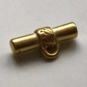 May include: A small, gold-coloured toggle clasp. The clasp has a cylindrical bar with rounded ends and a decorative, textured centre piece. A small loop is attached to the centre piece. The clasp is likely for jewellery or other small accessories.