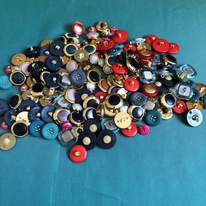 May include: A large collection of assorted buttons in various colours, shapes, and sizes. The buttons are made of different materials, including plastic, metal, and fabric. Some buttons have a shiny finish, while others have a matte finish.
