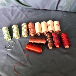 May include: A collection of spools of thread in various colors, including green, brown, and red. The spools are cylindrical and made of a light-colored material. Some threads are unwound, showing the thread's texture. The background is a dark blue fabric.