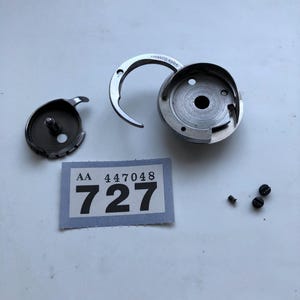 May include: A collection of sewing machine parts, including a bobbin case, hook, and screws. A label with the numbers "AA 447048 727" is also present. The parts are made of metal and are arranged on a light-colored surface.