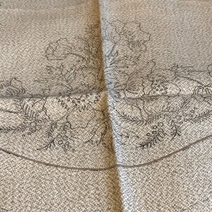 May include: A close-up of a beige fabric with a black embroidered design of flowers and leaves. The design is centred on the fabric and is surrounded by a border.