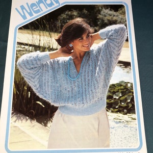 May include: A light blue knitted sweater with a ribbed pattern. The sweater has a V-neck and long sleeves. The model is wearing a white skirt and a blue necklace. The image is from a knitting pattern book titled "Wendy Dolce and Donna 2354". The sweater is for a bust size of 76-97 cm.