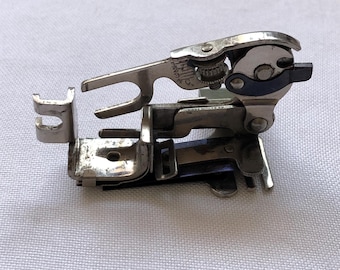 May include: A vintage silver sewing machine foot, featuring intricate mechanical details. The foot has a curved design with multiple levers and a small, dark blue accent. The metal shows signs of wear, indicating its age and use.