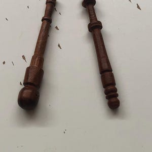 May include: Two dark brown wooden bobbins, likely for lacemaking, are displayed against a white surface. Each bobbin has a distinct shape, with a rounded or cylindrical body and a decorative top. The wood grain is visible.