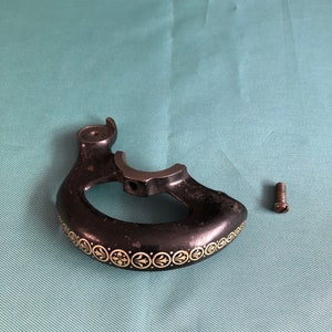 May include: Black metal sewing machine handle with a decorative cream-colored floral pattern along the edge. A small screw is next to the handle. The handle has a curved design with an open center.