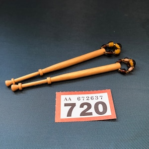 May include: Two light brown wooden lace bobbins, each with a decorative top of amber-coloured glass beads. They are set against a dark teal background. A small orange and white tag displays the number 720.