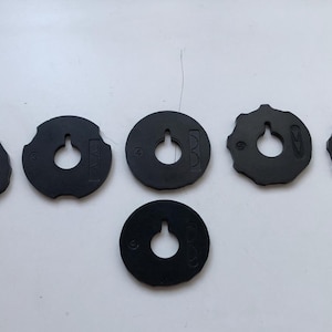 May include: A collection of black, circular objects with a hole in the center and a small notch at the top. Some have numbers or markings. The objects are arranged on a white surface.