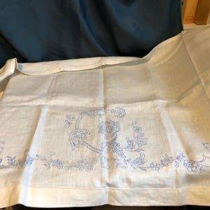 May include: A white linen tablecloth with blue embroidered floral designs. The embroidery features a large floral motif in the center and a border of smaller floral designs.