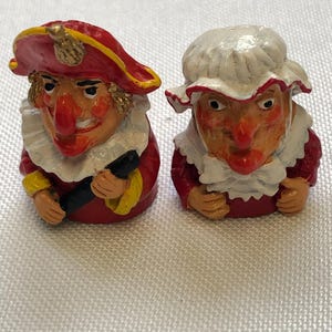 May include: Two small, hand-painted figurines. One figure wears a red coat and hat with gold trim, a white ruffled collar, and holds a black object. The other figure wears a red dress and a white bonnet with a ruffled collar.