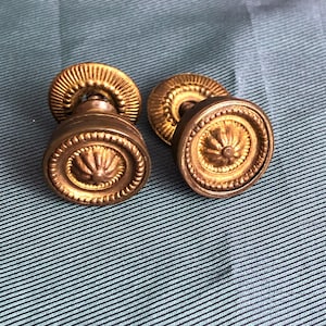 May include: Two antique gold-toned door knobs with a decorative floral design. The knobs are round and have a raised centre.