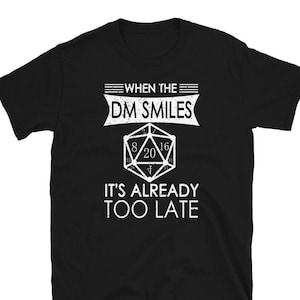 Puede incluir: Camiseta negra con texto blanco que dice "When the DM Smiles It's Already Too Late". Un dado blanco de 20 caras se presenta en el centro de la camiseta.