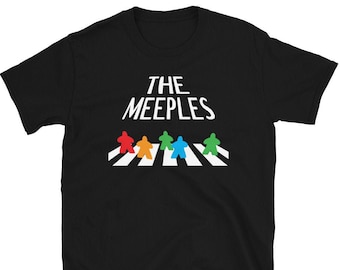 May include: Black t-shirt featuring the text "THE MEEPLES" in white, above a colorful graphic of meeple figures crossing a white and black crosswalk. The meeple figures are red, orange, green, and blue.