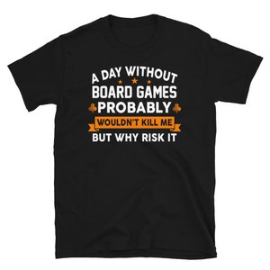 以下が含まれることがあります： 白い文字で「A day without board games probably wouldn't kill me but why risk it」と書かれた黒いTシャツ。テキストは星とクローバーのシンボルで囲まれています。