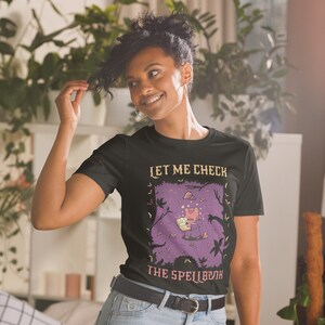 Puede incluir: Una camiseta negra con un gráfico morado y rosa de una rana sosteniendo un libro. El texto "Let me check the spellbook" está impreso en la camiseta.