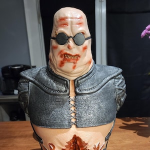 May include: A 3D printed bust of a character with a pale, fleshy complexion, wearing black sunglasses and a dark gray leather jacket with a laced front. The bust is mounted on a black base with a circular design.