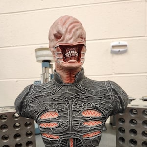 May include: A detailed bust of a creature with a grotesque, fleshy face and a mouth full of teeth. The creature is wearing a black, textured, armor-like garment with red accents. The bust is mounted on a red and gold base.