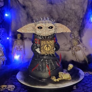 May include: A black and red detailed figure of a creature with large ears and a spiky head holding a box with a symbol. The figure is on a white circular platform with a black base. The figure is surrounded by a black and white background with a skeleton and a skull.