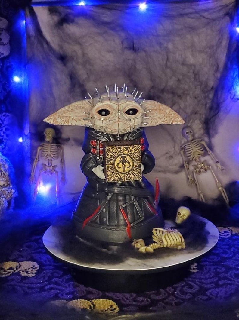 May include: A black and red detailed figure of a creature with large ears and a spiky head holding a box with a symbol. The figure is on a white circular platform with a black base. The figure is surrounded by a black and white background with a skeleton and a skull.