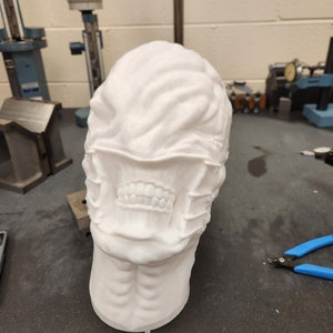 May include: A white, three-dimensional printed bust of a creature with a textured, brain-like cranium, exposed teeth, and a series of horizontal, stitched-like bands across the mid-face area, resting on a flat, dark gray industrial surface.