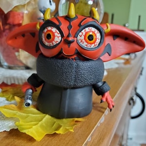 May include: A red and black Yoda figurine with horns and large eyes. The figurine is wearing a black coat and holding a lightsaber.