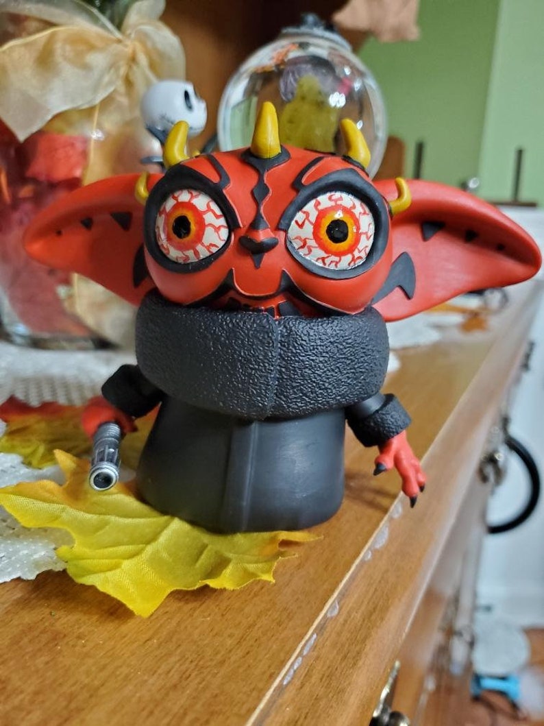 May include: A red and black Yoda figurine with horns and large eyes. The figurine is wearing a black coat and holding a lightsaber.