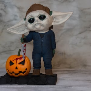 K&ouml;nnte beinhalten: Eine kleine, gr&uuml;nh&auml;utige Figur eines Charakters aus dem Star Wars-Franchise, gekleidet in einen blauen Overall und mit einem blutigen Messer in der Hand. Die Figur steht neben einem geschnitzten orangefarbenen K&uuml;rbis mit einem gruseligen Gesicht.