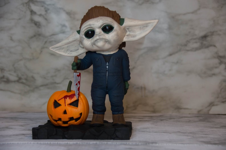 K&ouml;nnte beinhalten: Eine kleine, gr&uuml;nh&auml;utige Figur eines Charakters aus dem Star Wars-Franchise, gekleidet in einen blauen Overall und mit einem blutigen Messer in der Hand. Die Figur steht neben einem geschnitzten orangefarbenen K&uuml;rbis mit einem gruseligen Gesicht.