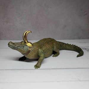 May include: A green and gold figurine of an alligator wearing a golden horned helmet and a brown strap. The alligator is on a white wooden surface with a gray background. The figurine has detailed scales and a realistic appearance.