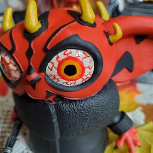 May include: A red and black toy figure with yellow horns and a large, white eye with red and orange details. The figure is wearing a black jacket.