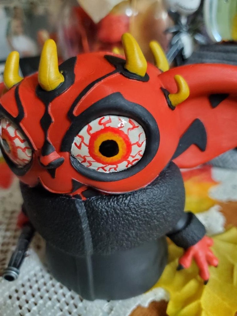 May include: A red and black toy figure with yellow horns and a large, white eye with red and orange details. The figure is wearing a black jacket.