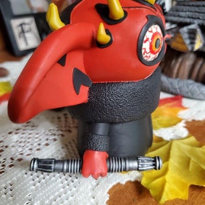 May include: A red and black Darth Maul action figure holding a silver lightsaber. The figure has a red face with a single yellow eye and horns.