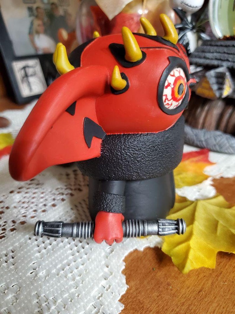 May include: A red and black Darth Maul action figure holding a silver lightsaber. The figure has a red face with a single yellow eye and horns.