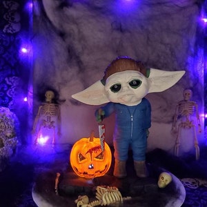 K&ouml;nnte beinhalten: Ein Halloween-Diorama mit einer kleinen, gr&uuml;nh&auml;utigen Figur mit gro&szlig;en Ohren, die ein blutiges Messer h&auml;lt. Die Figur steht vor einer beleuchteten K&uuml;rbislaterne und einem Skelett. Das Diorama ist vor einem dunklen, marmorierten Hintergrund platziert.