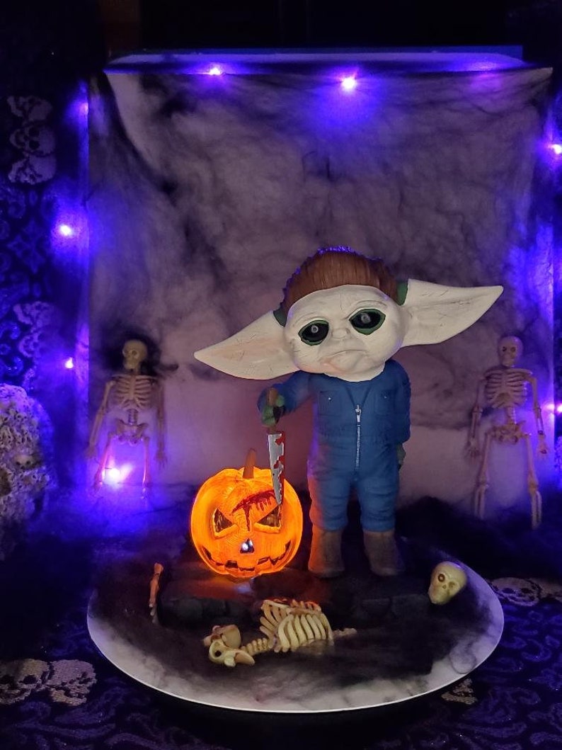 K&ouml;nnte beinhalten: Ein Halloween-Diorama mit einer kleinen, gr&uuml;nh&auml;utigen Figur mit gro&szlig;en Ohren, die ein blutiges Messer h&auml;lt. Die Figur steht vor einer beleuchteten K&uuml;rbislaterne und einem Skelett. Das Diorama ist vor einem dunklen, marmorierten Hintergrund platziert.