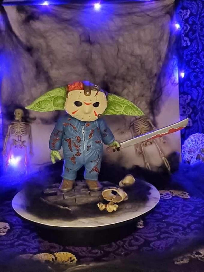 Puede incluir: Una figura verde y azul de un personaje que lleva una m&aacute;scara de hockey y sostiene un cuchillo ensangrentado. La figura est&aacute; de pie sobre una base redonda con un dise&ntilde;o en blanco y negro. La figura est&aacute; rodeada de un fondo oscuro con algunas luces.