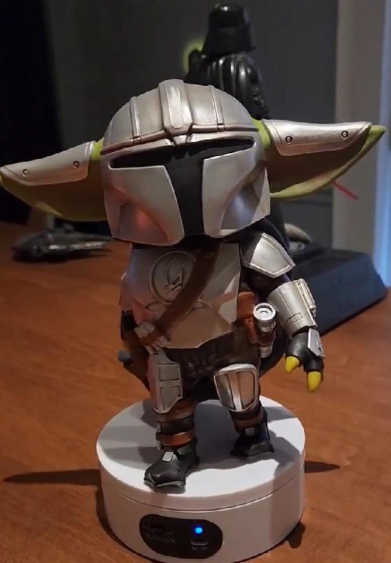 May include: A figurine of a character from The Mandalorian, featuring a silver helmet and armor, with large green ears and yellow hands. The figure is standing on a white rotating base, with a dark background.