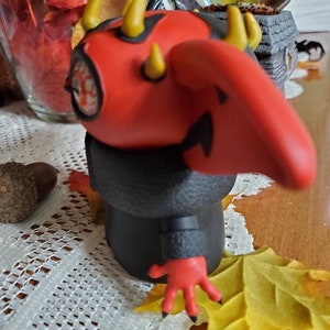May include: A red and black figurine with horns and a long nose. The figurine has a black coat and red hands. The figurine is standing on a table with a white tablecloth.