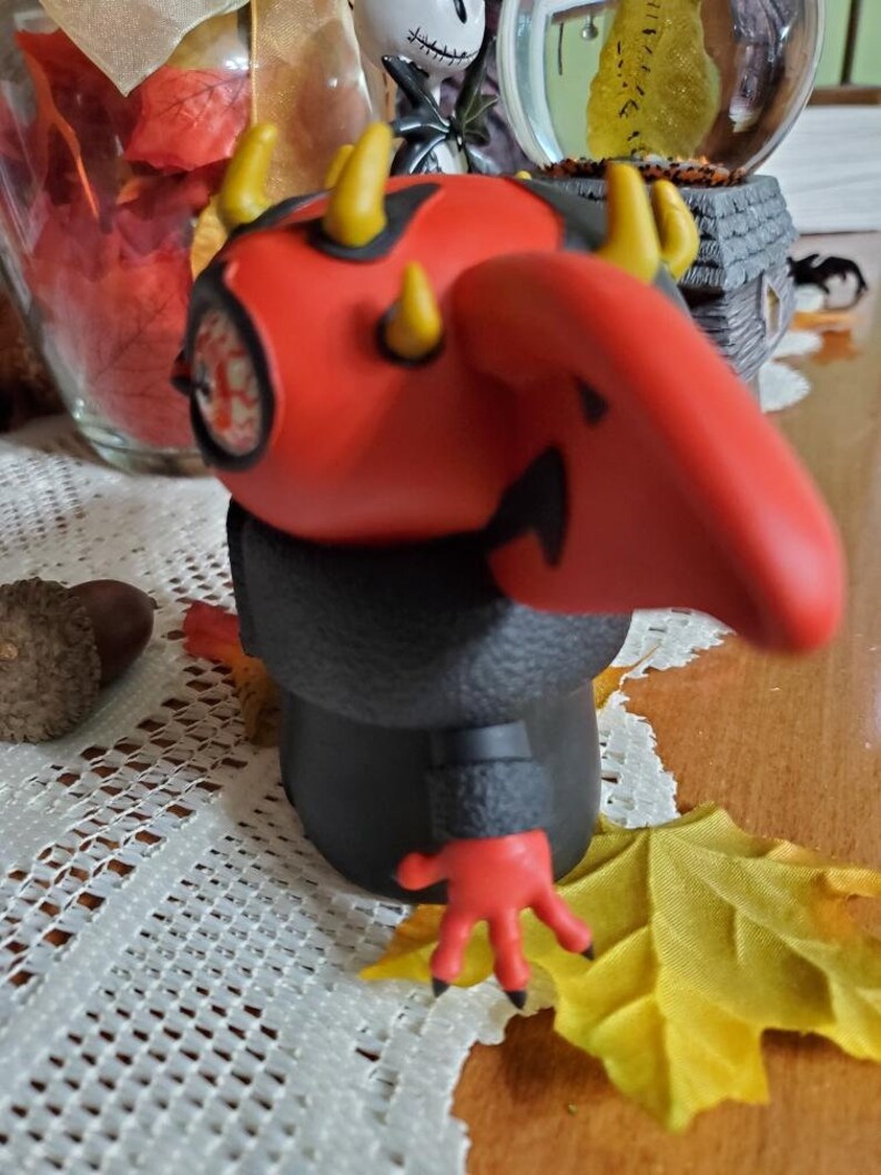 May include: A red and black figurine with horns and a long nose. The figurine has a black coat and red hands. The figurine is standing on a table with a white tablecloth.