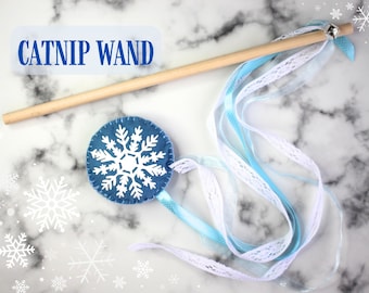 Pode incluir: Uma varinha de erva-cidreira de feltro azul com um desenho de floco de neve branco. A varinha tem um bastão de madeira e fitas azuis e brancas.