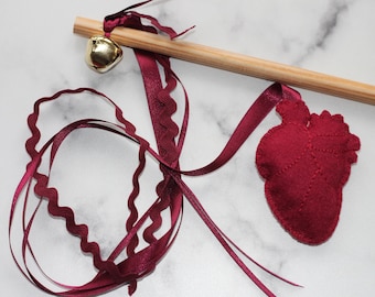 Puede incluir: Un juguete para gatos burdeos con una varilla de madera, una campana dorada y un corazón de fieltro. El juguete tiene cintas burdeos con borde festoneado y acabado satinado. El corazón de fieltro está cosido con un patrón de hojas.