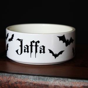 May include: A white ceramic bowl featuring the name Jaffa printed in a black gothic-style font on the exterior, surrounded by several black bat silhouettes. The bowl has a smooth, cylindrical shape with a clean, minimalist white interior finish.