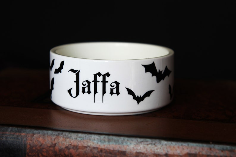 May include: A white ceramic bowl featuring the name Jaffa printed in a black gothic-style font on the exterior, surrounded by several black bat silhouettes. The bowl has a smooth, cylindrical shape with a clean, minimalist white interior finish.
