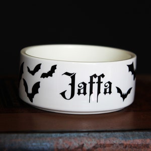 May include: A white ceramic bowl featuring black silhouettes of flying bats and the name Jaffa printed in a gothic-style black font on the exterior side. The bowl has a smooth, cylindrical shape with a clean white interior and a flat base.