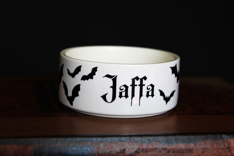May include: A white ceramic bowl featuring black silhouettes of flying bats and the name Jaffa printed in a gothic-style black font on the exterior side. The bowl has a smooth, cylindrical shape with a clean white interior and a flat base.
